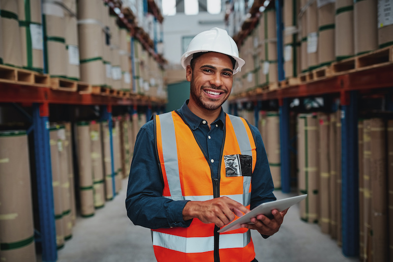 Happy male factory manager using digital tablet in warehouse while standing against goods shelf looking at camera man using manufacturing accounting software