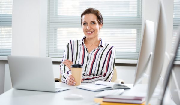 Business woman holding a coffee sitting at a laptop in bright office