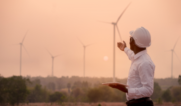 Man in hardhat outside gesturing at wind turbines during a hazy sunset