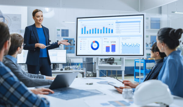 Modern Industrial Factory Meeting: Confident Female Engineer Uses Interactive Whiteboard, Makes Report to a Group of Engineers, Managers Talks and Shows Statistics, Growth and Analysis Information